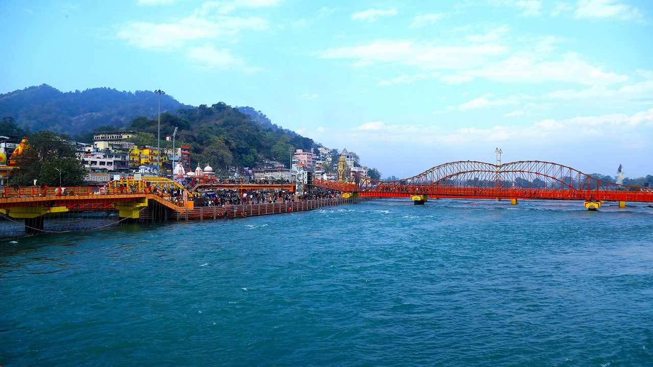 A scenic view of the Ganges River in Haridwar, India, with a crowd on riverbanks, colorful buildings, a red footbridge, and green hills under a partly cloudy sky.